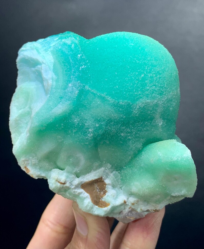 Puede incluir: Un vibrante esp&eacute;cimen mineral cristalino con un llamativo tono turquesa. El mineral tiene una forma redondeada y bulbosa con una superficie texturizada, y una base de color m&aacute;s claro, blanquecino y marr&oacute;n. La apariencia general es suave y pulida.