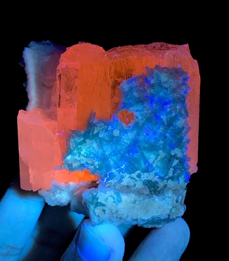 K&ouml;nnte beinhalten: Ein rohes Mineralexemplar mit starker Fluoreszenz unter ultraviolettem Licht. Die Kristallstruktur zeigt leuchtend orangefarbene Bereiche im Kontrast zu hellen, elektrisch blauen Kristallclustern auf einer texturierten, hellen Basis.