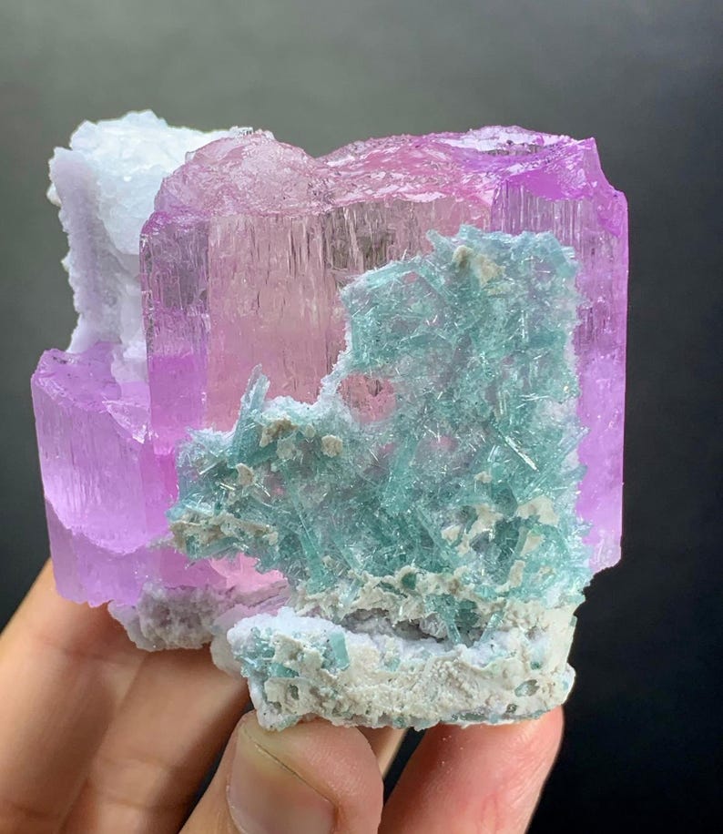 K&ouml;nnte beinhalten: Ein rohes Mineralexemplar mit einer gro&szlig;en, durchscheinenden rosa Kristallformation, die durch ein B&uuml;ndel leuchtend t&uuml;rkisfarbener, nadelartiger Kristallstrukturen kontrastiert wird. Das St&uuml;ck enth&auml;lt Bereiche aus wei&szlig;em, texturiertem Matrixgestein an der Basis.
