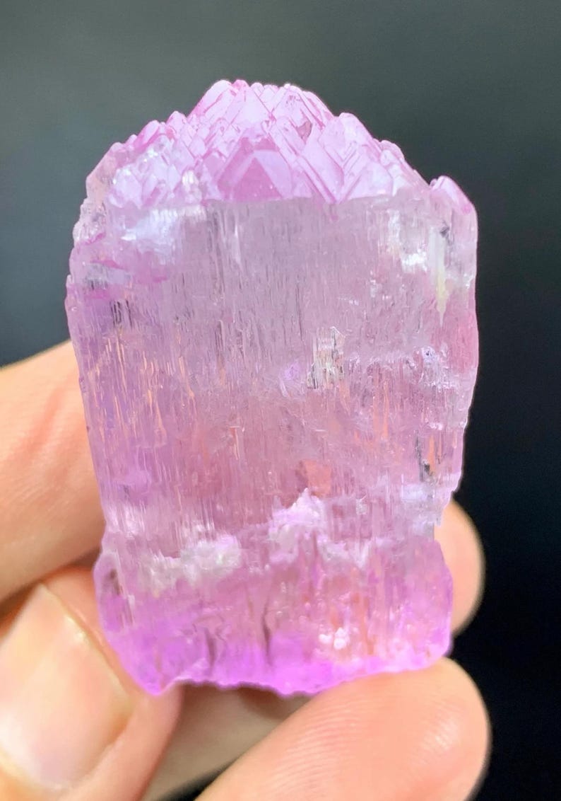 May include: A pink, translucent, faceted crystal with a striated surface. The crystal is approximately 2 inches tall and 1 inch wide.