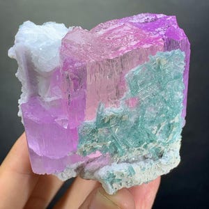 K&ouml;nnte beinhalten: Ein rohes Mineralexemplar mit gro&szlig;en, durchscheinenden, leuchtend rosa Kristallformationen. Die rosa Kristalle werden durch Ansammlungen von hellt&uuml;rkisfarbenen, nadelartigen Kristallstrukturen und eine texturierte, undurchsichtige wei&szlig;e Mineralbasis akzentuiert.