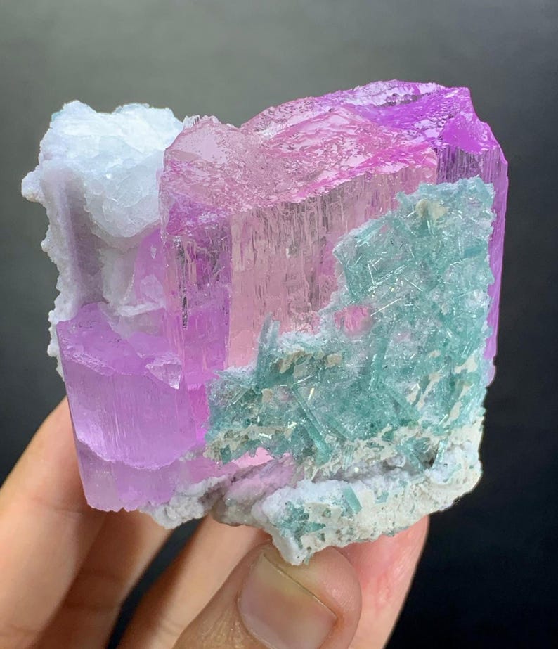 K&ouml;nnte beinhalten: Ein rohes Mineralexemplar mit gro&szlig;en, durchscheinenden, leuchtend rosa Kristallformationen. Die rosa Kristalle werden durch Ansammlungen von hellt&uuml;rkisfarbenen, nadelartigen Kristallstrukturen und eine texturierte, undurchsichtige wei&szlig;e Mineralbasis akzentuiert.