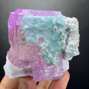 K&ouml;nnte beinhalten: Ein Mineralexemplar mit einer gro&szlig;en, durchscheinenden, hellvioletten Kristallbasis, auf der ein Cluster aus nadelartigen, hellblauen Kristallen w&auml;chst, akzentuiert durch Flecken aus undurchsichtigen, wei&szlig;en Mineralablagerungen.