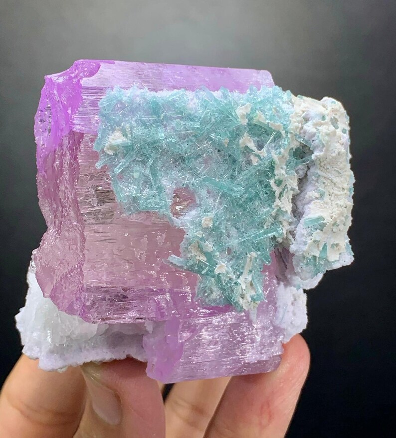 K&ouml;nnte beinhalten: Ein Mineralexemplar mit einer gro&szlig;en, durchscheinenden, hellvioletten Kristallbasis, auf der ein Cluster aus nadelartigen, hellblauen Kristallen w&auml;chst, akzentuiert durch Flecken aus undurchsichtigen, wei&szlig;en Mineralablagerungen.