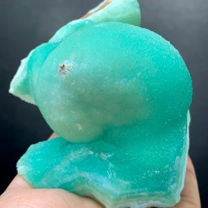 Puede incluir: Un vibrante esp&eacute;cimen mineral cristalino en tonos turquesa y verde claro. El mineral tiene una superficie redondeada y texturizada con una apariencia en capas en la base. El esp&eacute;cimen se sostiene en una mano, mostrando su belleza natural.