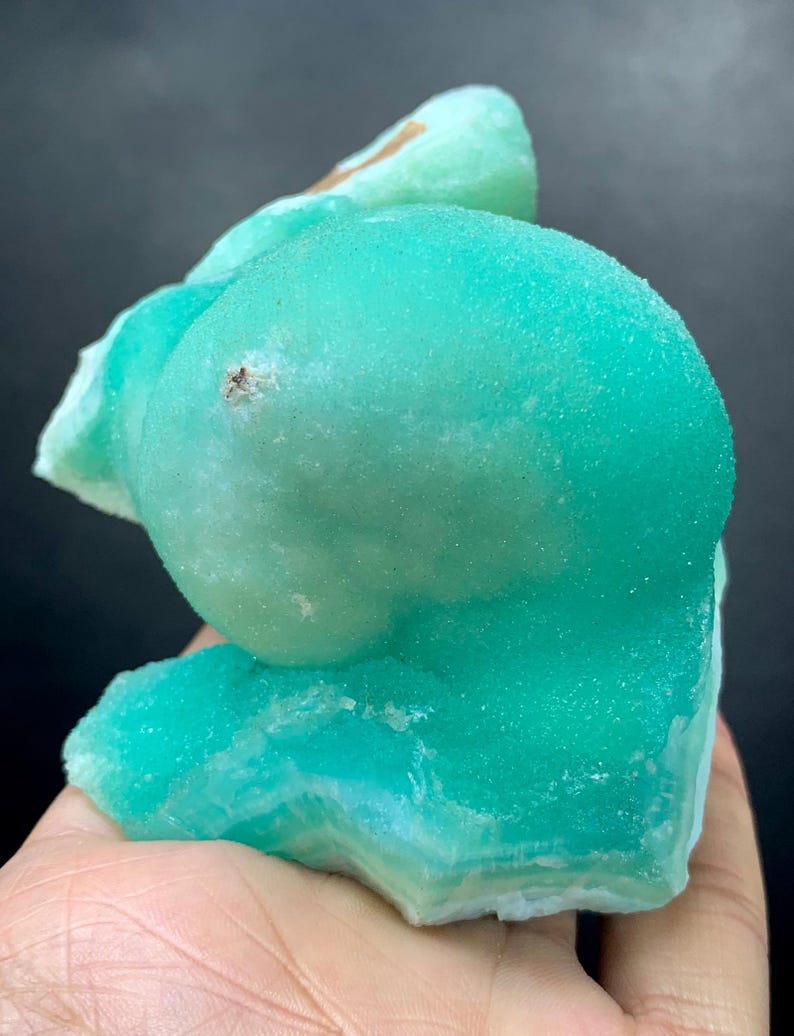 Puede incluir: Un vibrante esp&eacute;cimen mineral cristalino en tonos turquesa y verde claro. El mineral tiene una superficie redondeada y texturizada con una apariencia en capas en la base. El esp&eacute;cimen se sostiene en una mano, mostrando su belleza natural.