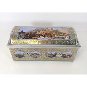 May include: A rectangular metal box with a curved lid. The lid features a colourful illustration of a townscape with the word "Nürnberg". The box is silver with gold filigree and additional townscape illustrations.