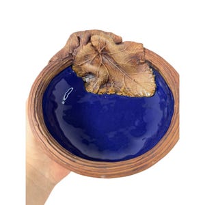 May include: A handcrafted ceramic bowl featuring a rich blue interior and a textured, brown exterior. A detailed, brown leaf design adorns the rim. The bowl is held by a hand against a white backdrop.