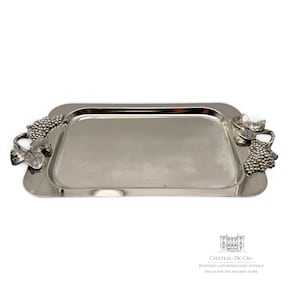 May include: A rectangular silver-toned metal serving tray featuring decorative handles shaped like grape clusters and leaves on both sides. The text CHATEAU DE CRU RESTORED AND REIMAGINED ANTIQUE PIECES FOR THE MODERN HOME is printed in the bottom right corner.
