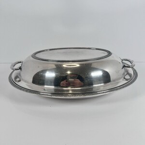 May include: A silver serving dish with a lid and two handles. The dish sits on a matching base with a decorative border. The reflective surface shows a distorted reflection of the surroundings.