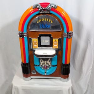 Puede incluir: Una gramola Crosley iJuke de estilo retro con un mueble de madera y detalles coloridos. La gramola presenta una parte superior cromada, un arco azul y naranja, y el texto "Crosley Juke" e "iJuke". La parte delantera tiene un área de altavoz azul y blanca.