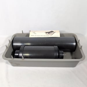 Könnte beinhalten: Eine graue Kunststoffschale mit zwei schwarzen zylindrischen Walzen und einer weißen Bedienungsanleitung mit der Aufschrift Detec Model 200 Color Print Processor. Die Schale hat kleine vertikale Kunststoffspitzen an den Innenkanten.