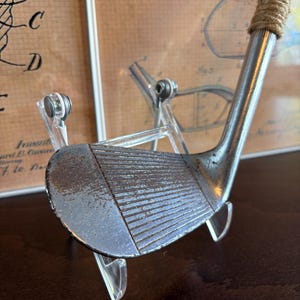 Puede incluir: Un palo de golf vintage con una cabeza de metal oxidada y una empu&ntilde;adura de cuero marr&oacute;n. El palo se muestra en un soporte de acr&iacute;lico transparente.