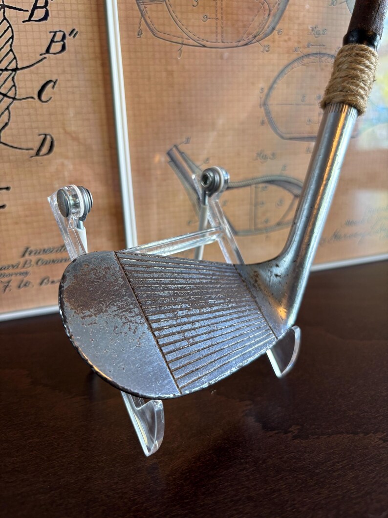 Puede incluir: Un palo de golf vintage con una cabeza de metal oxidada y una empu&ntilde;adura de cuero marr&oacute;n. El palo se muestra en un soporte de acr&iacute;lico transparente.
