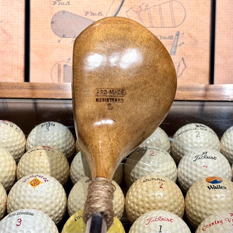 May include: A vintage wooden golf club with the text "PRO-MADE REGISTERED L" is displayed in front of a collection of golf balls. The club has a light brown finish and a wrapped grip. The golf balls are white with various brand names and numbers.