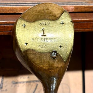 May include: Close-up of a vintage golf club head. The club head is made of wood and brass, with the number "1" and the word "REGISTERED" visible. The brass plate has the initials "H&B" and the number "4123".