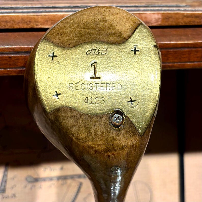 May include: Close-up of a vintage golf club head. The club head is made of wood and brass, with the number "1" and the word "REGISTERED" visible. The brass plate has the initials "H&B" and the number "4123".