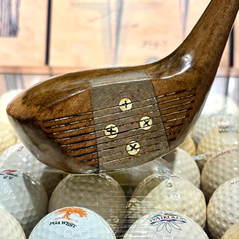 May include: A vintage wooden golf club with a dark brown head and a curved handle. The club head features a metal plate with three gold-colored markings. Golf balls are visible in the background.