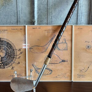 Puede incluir: Un palo de golf vintage de plata con una empu&ntilde;adura de cuero marr&oacute;n. El palo se muestra en un soporte de acr&iacute;lico transparente delante de tres dibujos de patentes enmarcados para equipos de golf.