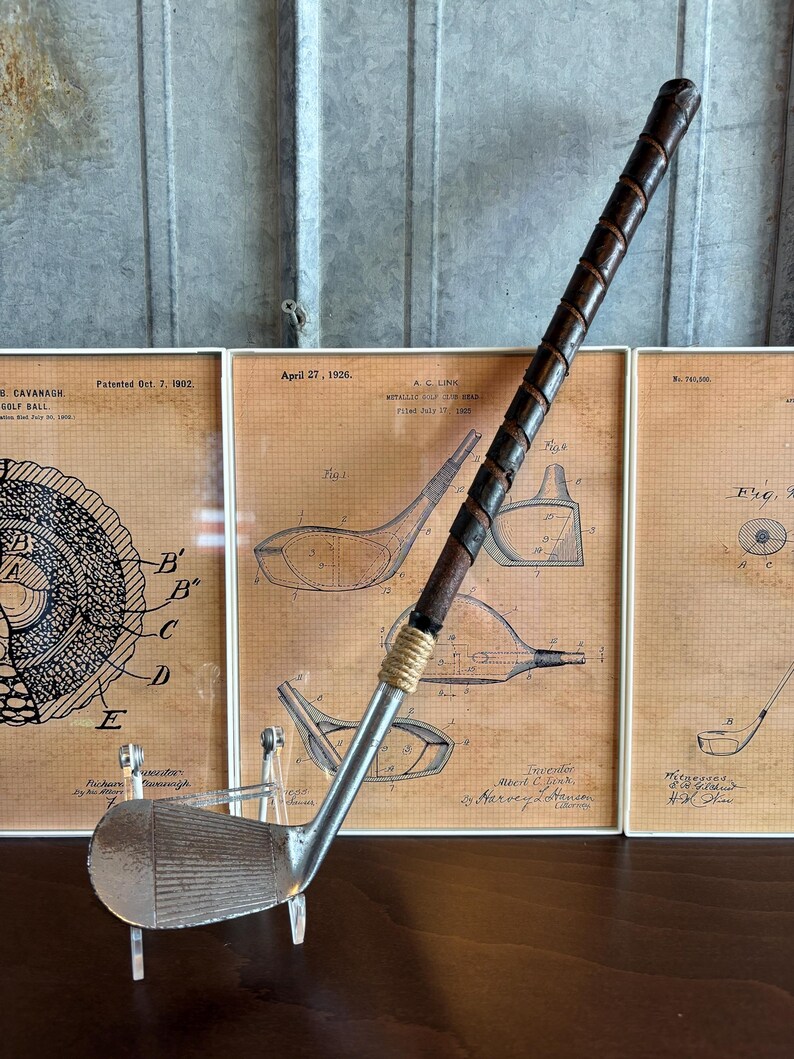 Puede incluir: Un palo de golf vintage de plata con una empu&ntilde;adura de cuero marr&oacute;n. El palo se muestra en un soporte de acr&iacute;lico transparente delante de tres dibujos de patentes enmarcados para equipos de golf.