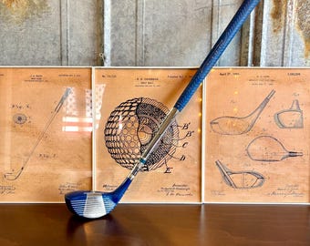 May include: A blue and silver golf club with a blue grip. The club is resting on a wooden surface in front of three framed vintage golf-related patent drawings.