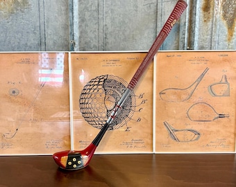 May include: A vintage wooden golf club with a red and black painted shaft and a black leather grip. The club is resting on a wooden table in front of three framed vintage golf patent drawings.