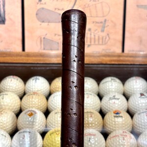 May include: Close-up of a vintage golf club handle with a dark brown, textured grip. The handle is positioned vertically in front of a collection of golf balls, some with visible logos. The background features diagrams.