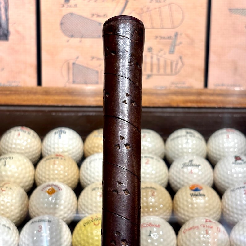 May include: Close-up of a vintage golf club handle with a dark brown, textured grip. The handle is positioned vertically in front of a collection of golf balls, some with visible logos. The background features diagrams.
