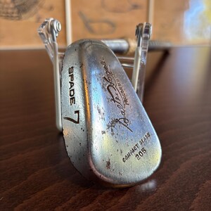 Puede incluir: Un palo de golf vintage de plata con el texto "SPADE 7" y "Balata-Flex" impreso en la cara. El palo tiene una etiqueta "Compact Blade" y el n&uacute;mero "205" impreso en la parte posterior.