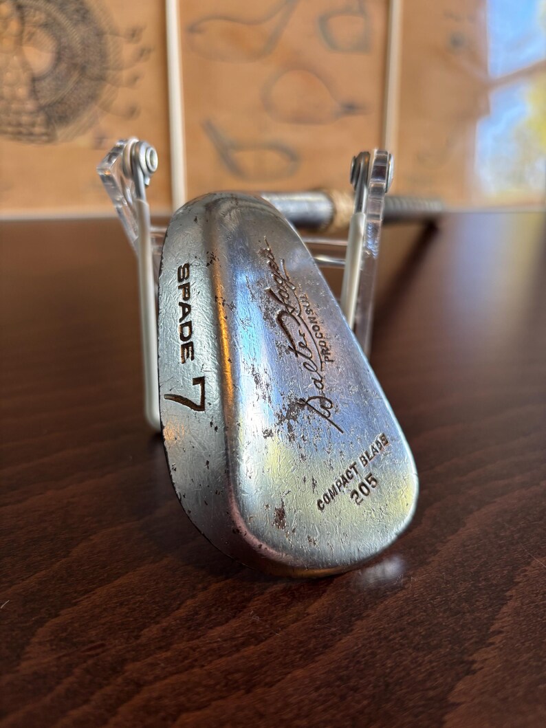 Puede incluir: Un palo de golf vintage de plata con el texto "SPADE 7" y "Balata-Flex" impreso en la cara. El palo tiene una etiqueta "Compact Blade" y el n&uacute;mero "205" impreso en la parte posterior.