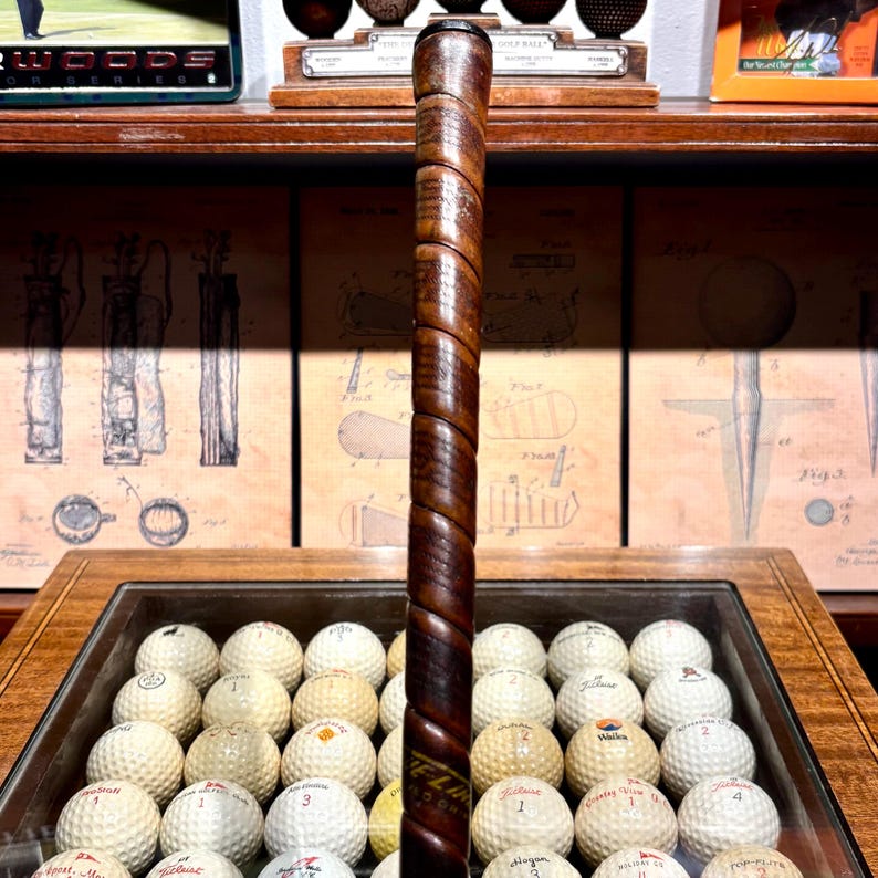 May include: A vintage golf club with a dark brown, spiraled leather grip. The club is positioned in front of a display case filled with golf balls. The background includes framed golf-related artwork and memorabilia.