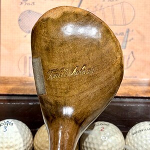 May include: A vintage wooden golf club with a light brown finish. The club head features the engraved name "Tom Nicholson" in a gold color. The club is positioned above golf balls, suggesting a golf-related theme.