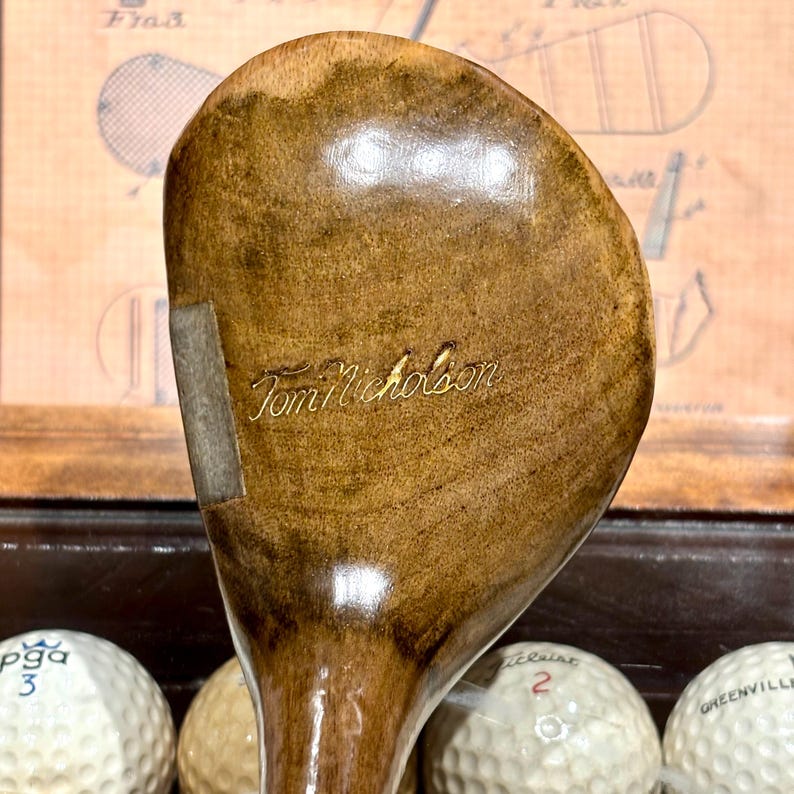 May include: A vintage wooden golf club with a light brown finish. The club head features the engraved name "Tom Nicholson" in a gold color. The club is positioned above golf balls, suggesting a golf-related theme.