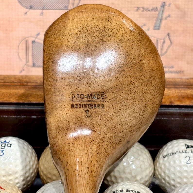 May include: A vintage wooden golf club with the text "PRO-MADE REGISTERED" stamped on the club head. The club has a light brown finish and is surrounded by golf balls. The club is a classic piece of sports equipment.