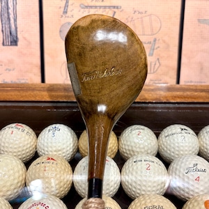 May include: A vintage wooden golf club with the name "Tom Nicholson" inscribed on the club head. The club has a dark brown finish and is surrounded by golf balls. The golf balls have various markings and numbers.