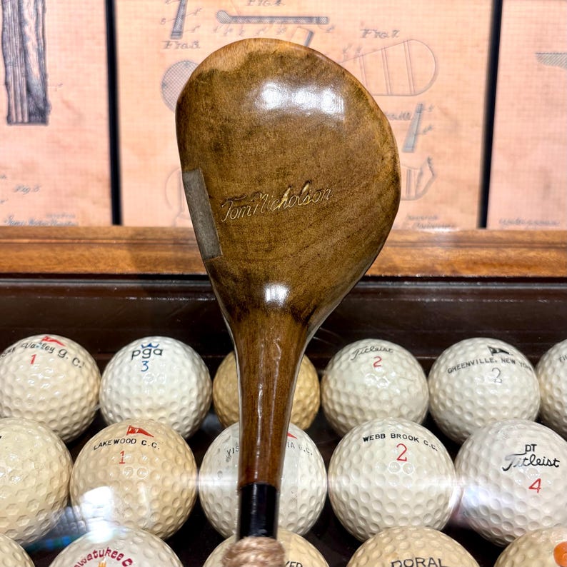 May include: A vintage wooden golf club with the name "Tom Nicholson" inscribed on the club head. The club has a dark brown finish and is surrounded by golf balls. The golf balls have various markings and numbers.