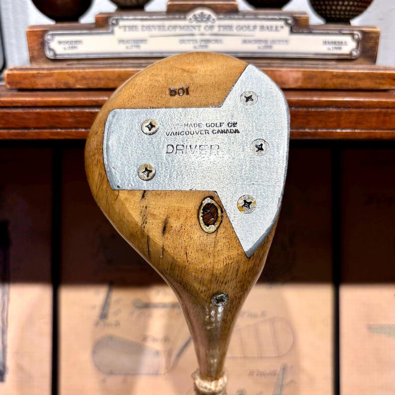May include: Vintage wooden golf driver with a silver-colored metal faceplate. The faceplate is secured with four screws and features the text "MADE GOLF OF VANCOUVER CANADA DRIVER". The club head is a light brown color.