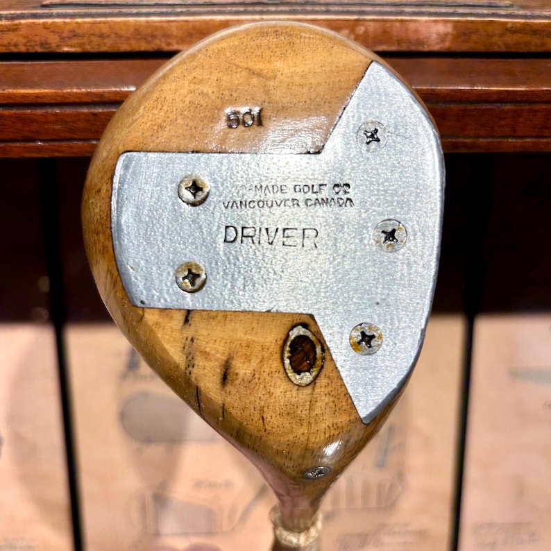 May include: Vintage wooden golf driver with a silver metal faceplate. The faceplate is secured with four screws and features the text "MADE GOLF CO VANCOUVER CANADA DRIVER". The club head is a light brown color.