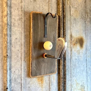 May include: A rustic golf club and ball display. The display features a dark gray wooden plaque with a rope border, a black metal hook, a golf ball, and a golf club. The golf club is silver and has the words "Ben Hogan" on it.