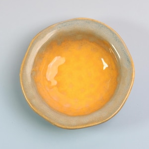 May include: A small, handmade ceramic bowl with a unique, organic shape. The interior is a vibrant yellow, transitioning to a golden hue towards the center. The rim has a muted blue-gray glaze, creating a striking contrast. The bowl is set against a light blue background.