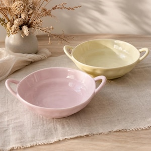 May include: Two ceramic bowls with handles, one in a soft pastel pink and the other in a pale yellow. Each bowl features a smooth, glossy finish and a rounded shape with two side handles for easy gripping, presented as a pair of kitchen serving dishes.