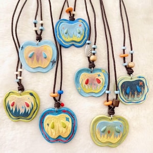May include: A collection of seven apple-shaped pendants on brown braided cords. Each pendant features a unique painted design with stylized flames, teardrop shapes, and small star accents. The necklaces are adorned with various colorful beads.