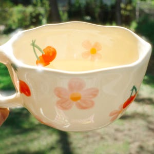 Könnte beinhalten: Cremefarbene Keramiktasse mit gewelltem Rand, verziert mit rosa Blumen und roten Kirschen. Die Tasse ist mit einer hellen Flüssigkeit gefüllt. Das Design ist floral und wird von einer Hand gehalten, was auf ein Getränk hindeutet.