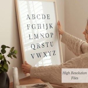 May include: A framed alphabet print in a light wood frame. The black letters are arranged in rows on a white background. The print is being held by a person. The text "High Resolution Files" is in the bottom right corner.