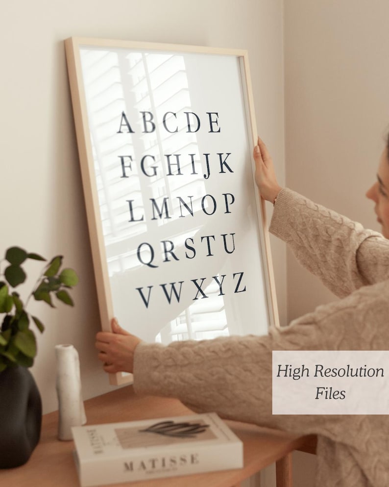 May include: A framed alphabet print in a light wood frame. The black letters are arranged in rows on a white background. The print is being held by a person. The text "High Resolution Files" is in the bottom right corner.