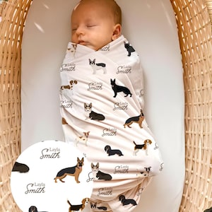 May include: A sleeping baby wrapped in a cream-colored swaddle blanket. The blanket is patterned with various dog breeds and the name "Layla Smith" in a cursive font. The baby is in a woven basket.