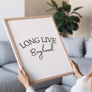 May include: A framed print with the words "LONG LIVE Boyhood" in black script. The print has a white background and a light wood frame. The image is held by a person in a light-colored sweater.