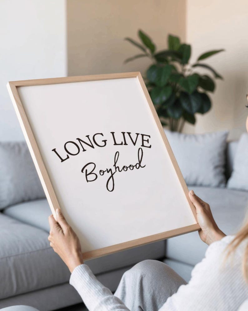 May include: A framed print with the words "LONG LIVE Boyhood" in black script. The print has a white background and a light wood frame. The image is held by a person in a light-colored sweater.