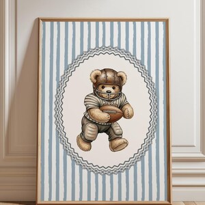 May include: A framed print featuring a teddy bear in a football uniform, including a helmet and pads, holding a football. The bear is set against a light blue and white striped background, framed by a decorative oval border.