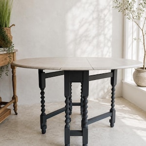 May include: A round, light-coloured wooden drop-leaf table with a dark grey base. The table has turned legs and a split tabletop. The table is approximately 122 cm in diameter when fully extended.